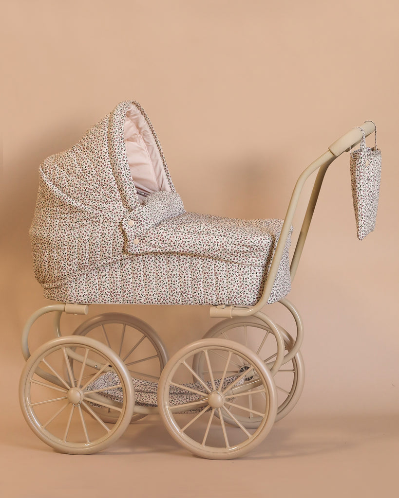 The Konges Sloejd Mini Doll Pram - Milk Tank is a vintage-style baby pram featuring quilted floral fabric, a large hood, and a matching bag that hangs on the handle. It has four large wheels and a light beige frame, complemented by a cushioned storage basket set against a plain background.