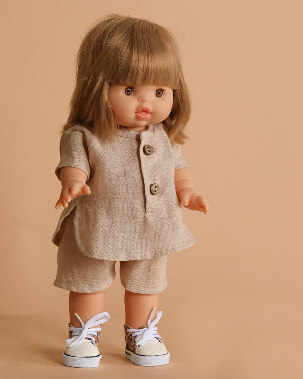 The Minikane Standing Doll (14") - Zoelie features straight blonde hair with bangs and stands against a plain beige background, dressed in a beige linen shorts set with two buttons and white sneakers with animal print details.