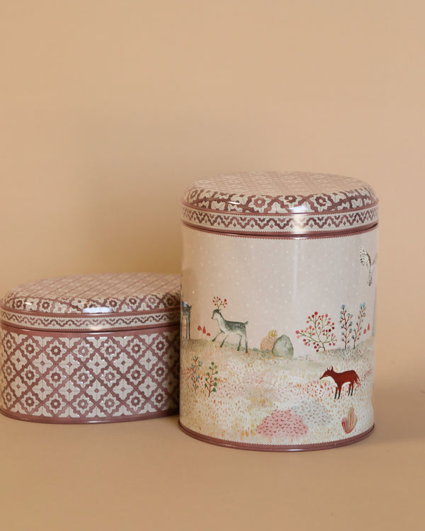 Displayed against a beige background are two decorative tins with lids from the Maileg Metal Box, 2 Piece Set - Winter Wonderland. The taller tin showcases a gentle nature scene with deer, foxes, and trees, while the shorter one displays a geometric pattern. These vintage-inspired tins are ideal for storing Christmas sweets.