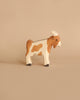 A small wooden figurine of a brown and white cow with black hooves is placed against a plain beige background. The Holztiger Billy-Goat, handcrafted out of wood, has a simple appearance with minimalistic features and a smooth finish.