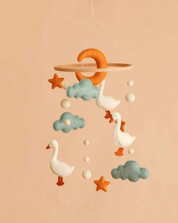 Handmade Mobile - The Sleepy Goose - Final Sale features felt ducks, clouds, stars, balls, and a crescent moon delicately hanging from a wooden hoop against a peach background. This charming nursery mobile adds a whimsical touch to any room.