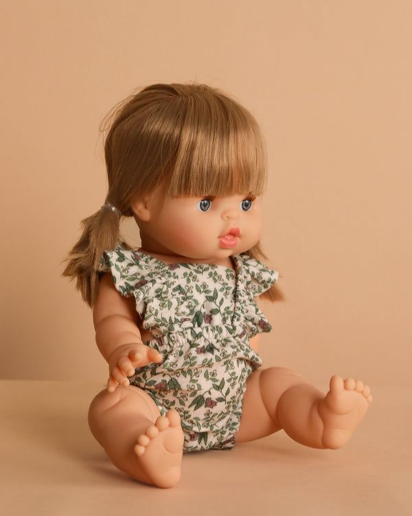 The Minikane Baby Doll (13") - Yze With Outfit, with blonde pigtails and a floral romper, sits on a beige surface. This anatomically correct doll features a natural vanilla scent.