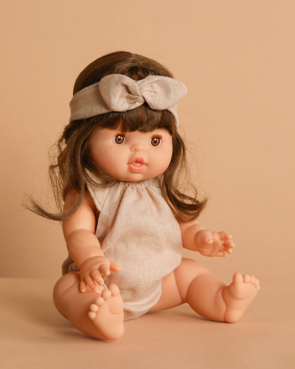 The Minikane Baby Doll (13") - Valentine With Outfit has brown hair, a beige bow headband, and a matching sleeveless romper. Seated on a beige surface with a beige background, this anatomically correct doll features a delicate vanilla scent.