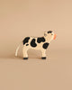 From the Holztiger Handmade Wooden Farm Animals Set, this wooden cow toy with black spots, an orange snout, and a white rope tail stands on a beige surface against a matching beige background.