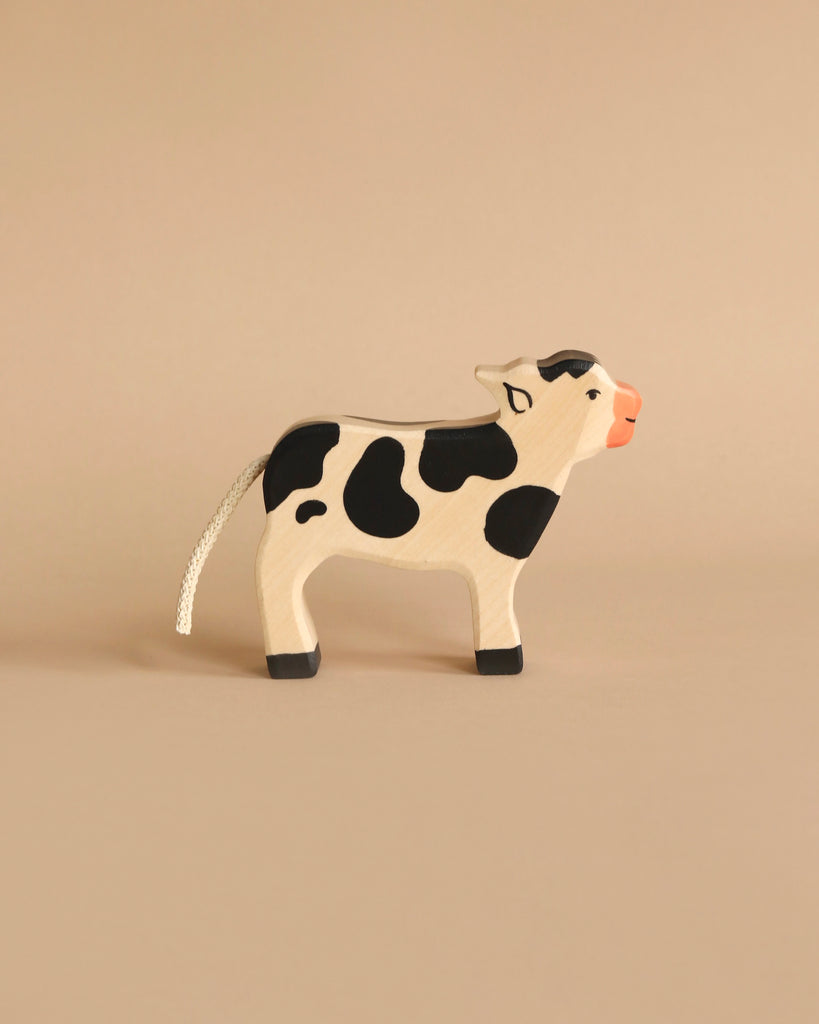 From the Holztiger Handmade Wooden Farm Animals Set, this wooden cow toy with black spots, an orange snout, and a white rope tail stands on a beige surface against a matching beige background.