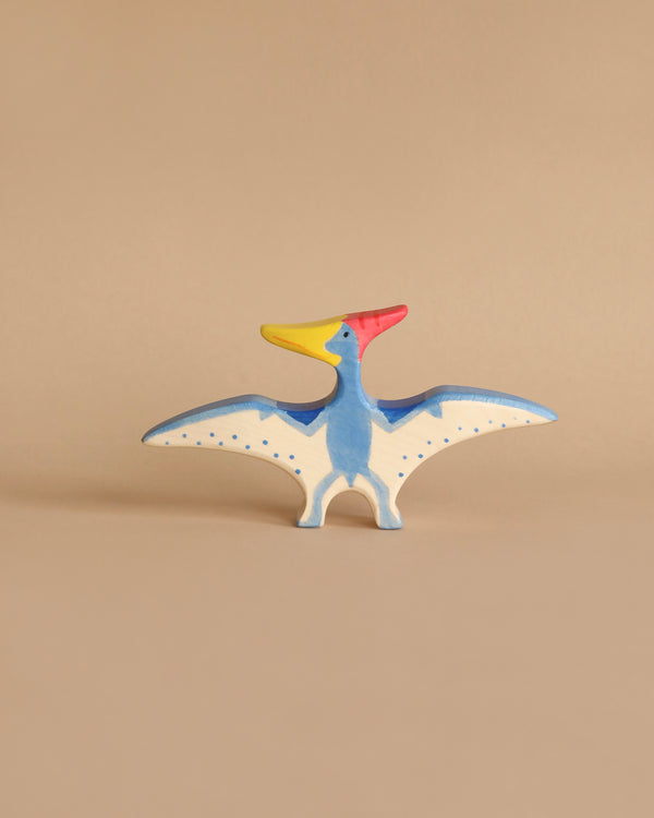 A small, colorful wooden toy figurine of a pteranodon with blue wings, a red crest, a yellow beak, and white underwings decorated with blue dots, stands against a plain beige background. Handcrafted out of maple and beech wood, this Holztiger Pteranodon Dinosaur is made in Europe.