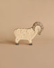 A small wooden toy resembling a ram stands on a neutral beige background. The Holztiger Ram features carved details, including a textured body, curved horns, and gray hooves. Its simple, minimalist design creates a charming appearance typical of handcrafted wood toys made in Europe.