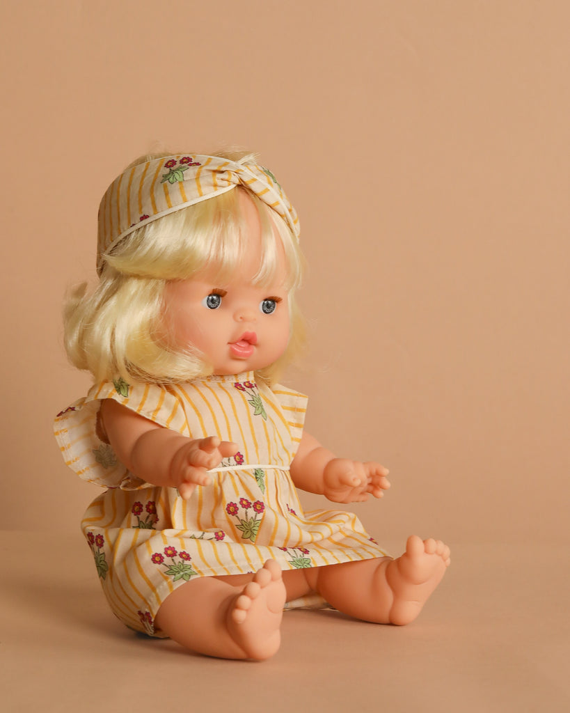 The Minikane Baby Doll (13") – Angela With Outfit features blonde hair, blue eyes, and a natural vanilla scent. She wears a cherry-patterned yellow-and-white striped dress with a matching headband and sits on a beige surface.