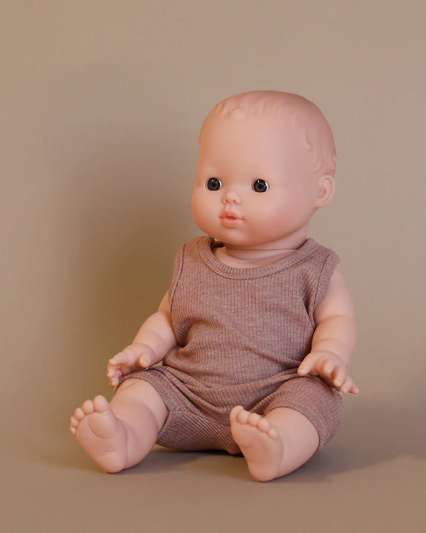 The Minikane Doll (13") - Louis with Clothing Set, crafted from phthalate-free vinyl, is displayed on a smooth surface with its legs extended and hands resting on its knees. Dressed in a sleeveless brown outfit, this realistic baby doll invites imaginative playtime against a plain beige background.