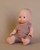 The Minikane Doll (13") - Louis with Clothing Set, crafted from phthalate-free vinyl, is displayed on a smooth surface with its legs extended and hands resting on its knees. Dressed in a sleeveless brown outfit, this realistic baby doll invites imaginative playtime against a plain beige background.