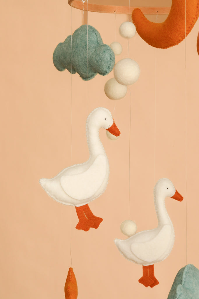 Two white felt duck ornaments hang from strings, surrounded by a blue felt cloud, orange accents, and small white felt balls against a peach background in "Handmade Mobile - The Sleepy Goose - Final Sale." Perfect as a decorative ceiling mobile, it adds a whimsical touch to any nursery collection.