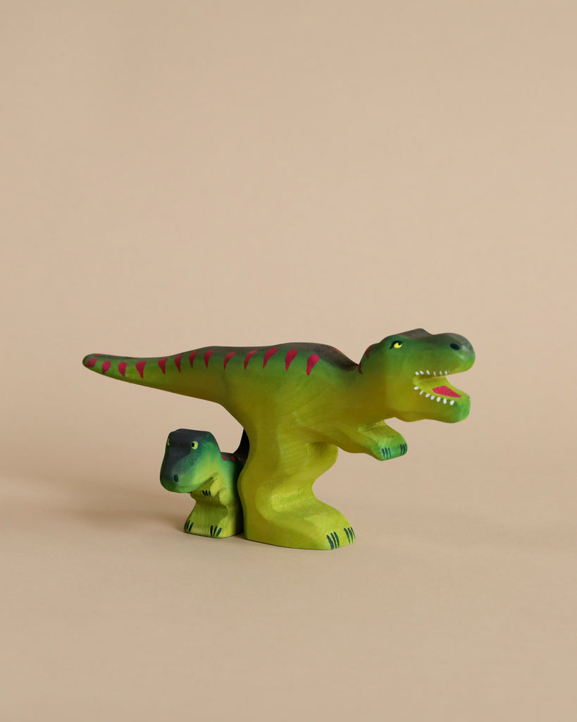 Two green Handmade Wooden T-Rex Dinosaur figurines, one large and one small with pink stripes, sit side by side on a beige background. The larger dinosaur's open mouth shows white teeth—ideal for the Easter Basket Gift Set.