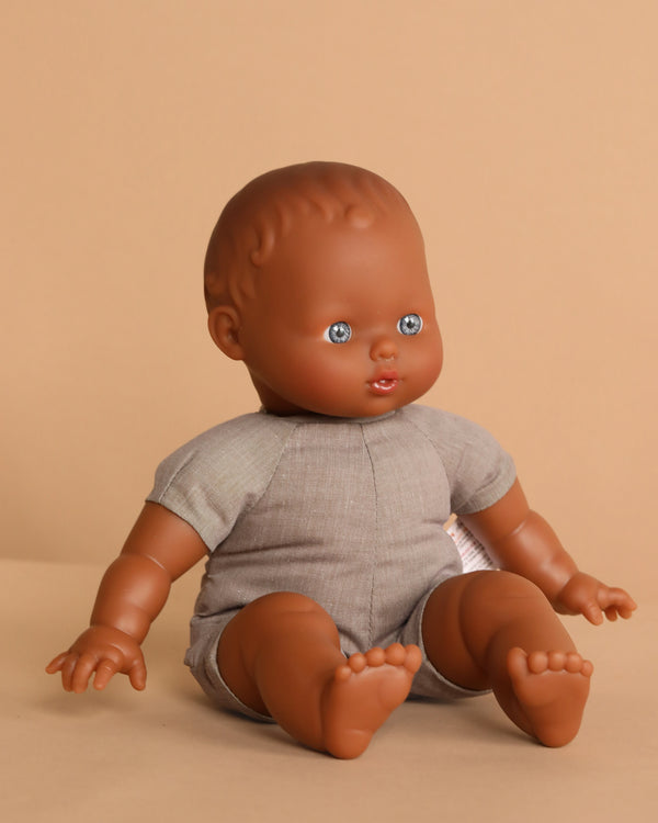 The Minikane Soft Body Doll (11”) - Suzie features curly textured hair, blue eyes, an open mouth, and a light gray romper. Crafted from phthalate-free vinyl, Suzie inspires imaginative play as she sits upright on a beige background.