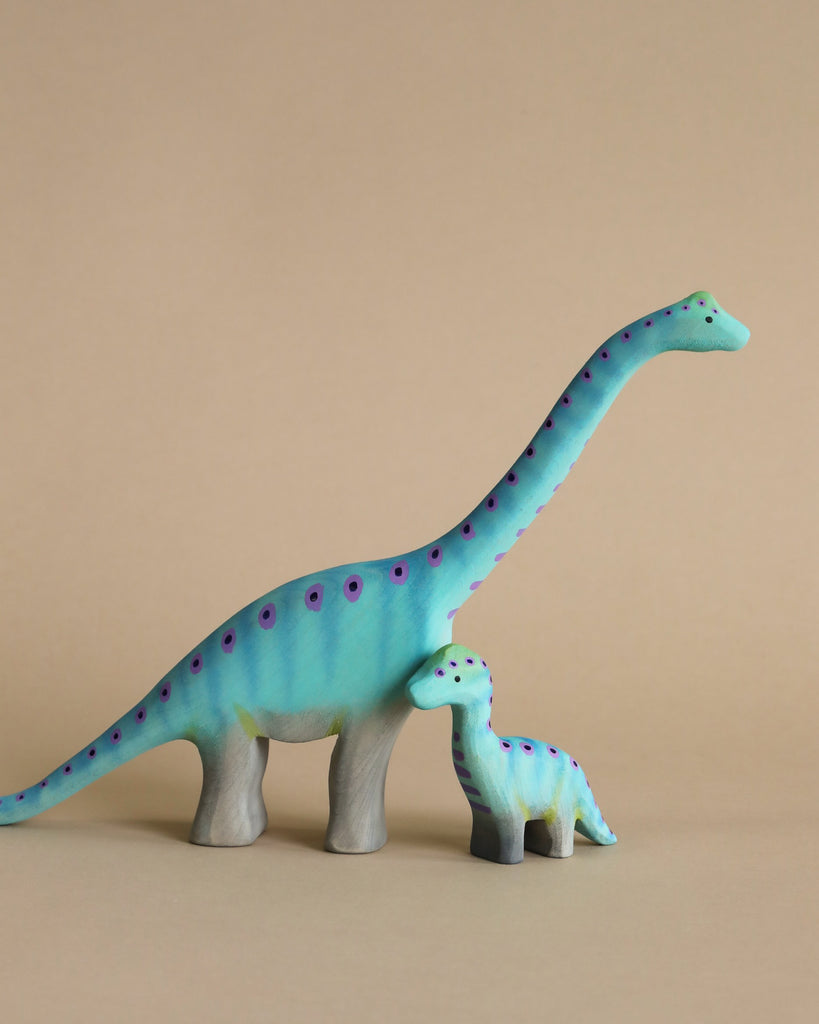 The Bumbu Handmade Wooden Prehistoric Set features two blue, purple-spotted handmade wooden dinosaurs—one large, one small—standing side by side. Painted with safe, non-toxic colors, this set inspires imaginative play on a beige background.
