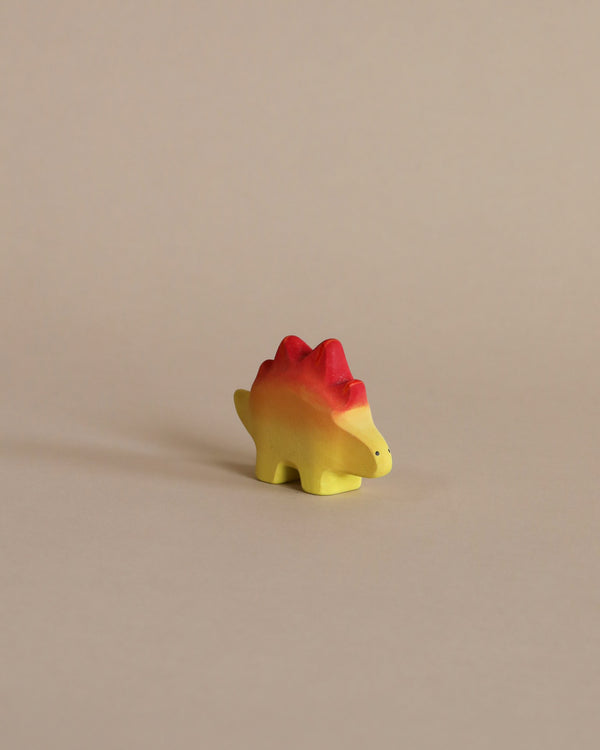 A small, colorful Handmade Stegosaurus Baby Dinosaur toy with a yellow body and a red-tipped back, shown in profile against a plain beige background.