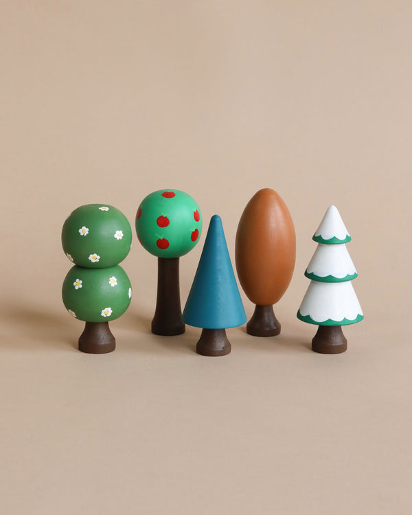 The Handmade Wooden Trees set features five beautifully hand-painted wooden toy trees, each with simple and colorful designs: a tree adorned with white flowers, one bearing red apples, a blue triangular tree, an oval-shaped brown tree, and a snow-covered tree. All are displayed side by side on a beige background.