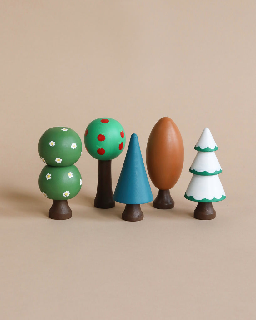 The Handmade Wooden Trees set features five beautifully hand-painted wooden toy trees, each with simple and colorful designs: a tree adorned with white flowers, one bearing red apples, a blue triangular tree, an oval-shaped brown tree, and a snow-covered tree. All are displayed side by side on a beige background.