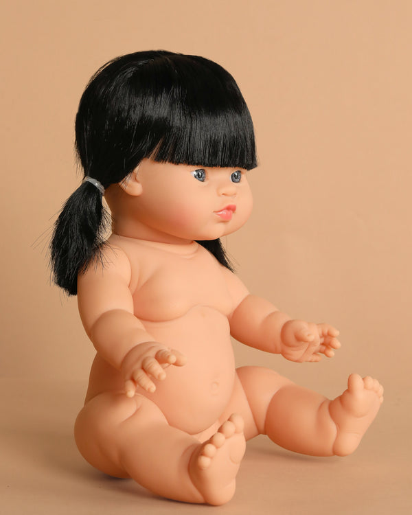 The Minikane Baby Doll (13") - Kaya, with black hair in pigtails, sits upright on a beige surface against a matching background. This anatomically correct collectible is pictured unclothed; clothing is sold separately.