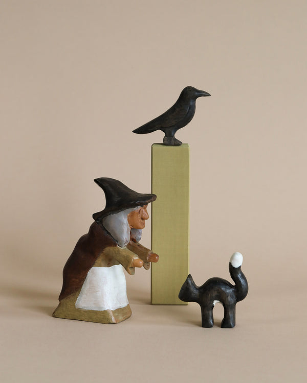 A figurine of The Witch and Her Cat & Crow, with a crow perched atop a tall, thin pedestal, all set against a light beige background.