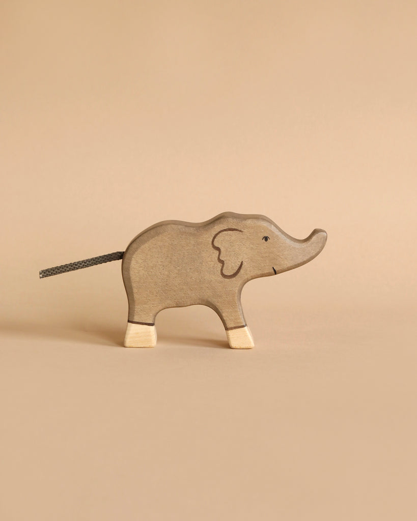 From the Holztiger Handmade Wooden Safari Animals Set, a light brown wooden elephant toy with white feet, a painted smile, and a dark cord tail stands against a beige background in simple, minimalist style.