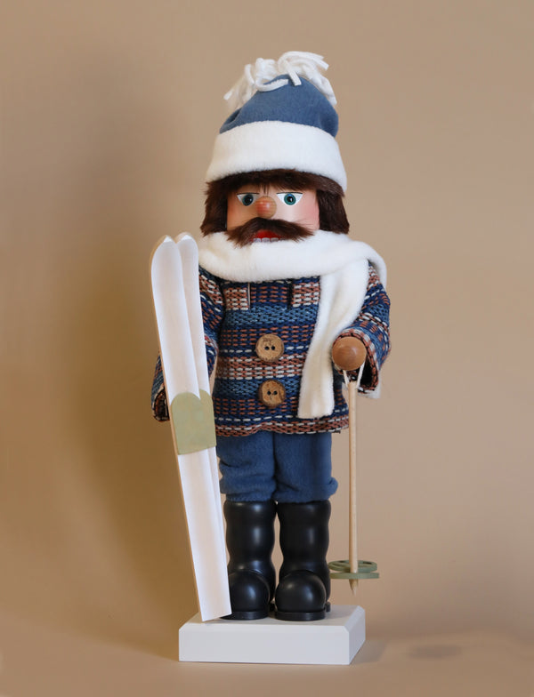 The Christian Ulbricht Collectible Wooden Nutcracker - Skier is expertly designed as a skier, featuring a blue and brown plaid coat, blue pants, and a blue hat. It holds white skis and ski poles, and it is highlighted by a white scarf and black boots against a beige backdrop.