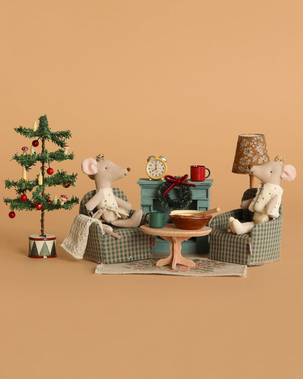 The Maileg Royal Twins Cozy Christmas Living Room Set features two royal mice on plaid armchairs, gathered around a table with festive accessories, a Christmas tree, and a lamp, creating a charming and cosy holiday scene.