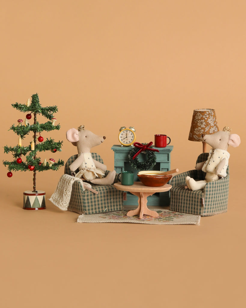 The Maileg Royal Twins Cozy Christmas Living Room Set features two royal mice on plaid armchairs, gathered around a table with festive accessories, a Christmas tree, and a lamp, creating a charming and cosy holiday scene.