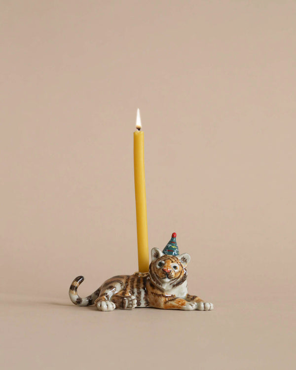 A Tiger Cake Topper with a party hat lying down supports a tall, lit yellow candle on its back against a soft pink background.