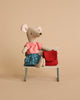 A Maileg Big Sister with Backpack - Red doll dressed in a red and white gingham shirt and blue plaid skirt sits on a miniature chair with a tiny red bag beside it, against a beige background.