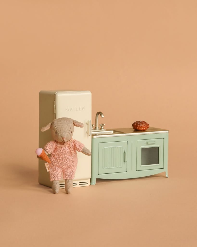 A small stuffed animal in a pink outfit holds an ice cream cone beside the Maileg Little Lambs Kitchen Set and Maileg Metal Fridge, with a pastry on the counter. The kitchen features a sink and oven against a beige background.