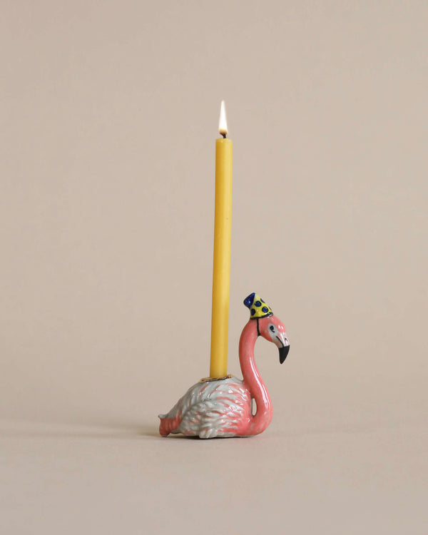 A ceramic Flamingo "Party Animal" Cake Topper candle holder, hand painted, with a party hat, supporting a lit yellow taper candle, set against a plain beige background.