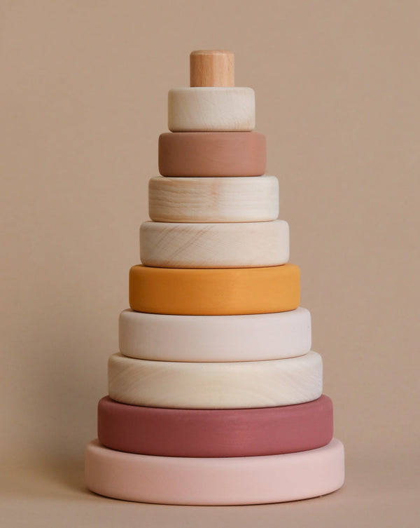 A wooden pyramid stacker toy with rings in pastel shades of pink, cream, yellow, and brown, designed to enhance hand-eye coordination, standing against a neutral beige background.