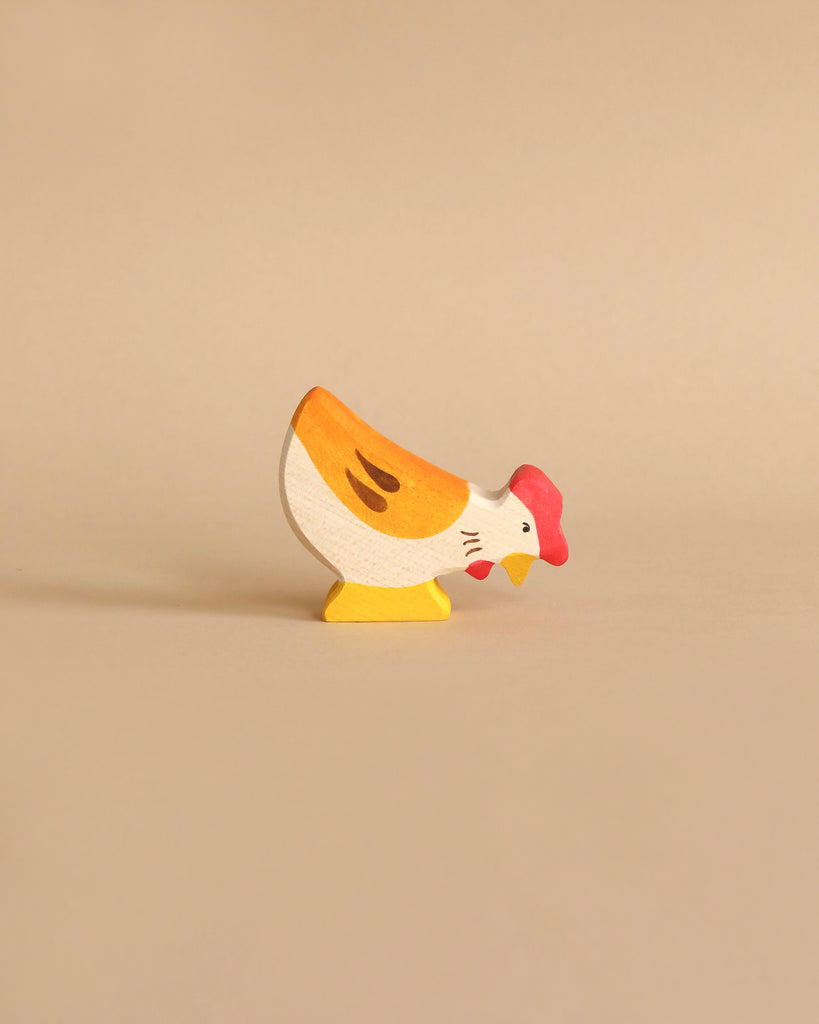 A small, hand-painted chicken from the Holztiger Handmade Wooden Animal Play Set, featuring a yellow body, red comb, and orange accents, stands on a beige background—highlighting European craftsmanship.