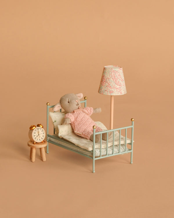 A small stuffed mouse in a pink sweater lies in the Maileg Little Lambs Bedroom Set, featuring a vintage-style bed with pillows and blanket, plus matching accessories: a nightstand with alarm clock and floor lamp. The background is tan.