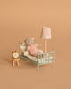A small stuffed mouse in a pink sweater lies in the Maileg Little Lambs Bedroom Set, featuring a vintage-style bed with pillows and blanket, plus matching accessories: a nightstand with alarm clock and floor lamp. The background is tan.