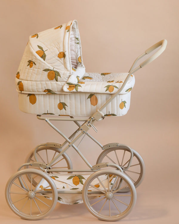 The Doll Pram - Grand Lemon is a vintage-style pram made from recycled materials, featuring a white quilted fabric adorned with a delightful lemon pattern. It includes a curved handle, large spoked wheels, and an interior made of plush organic cotton, all set against a neutral background.