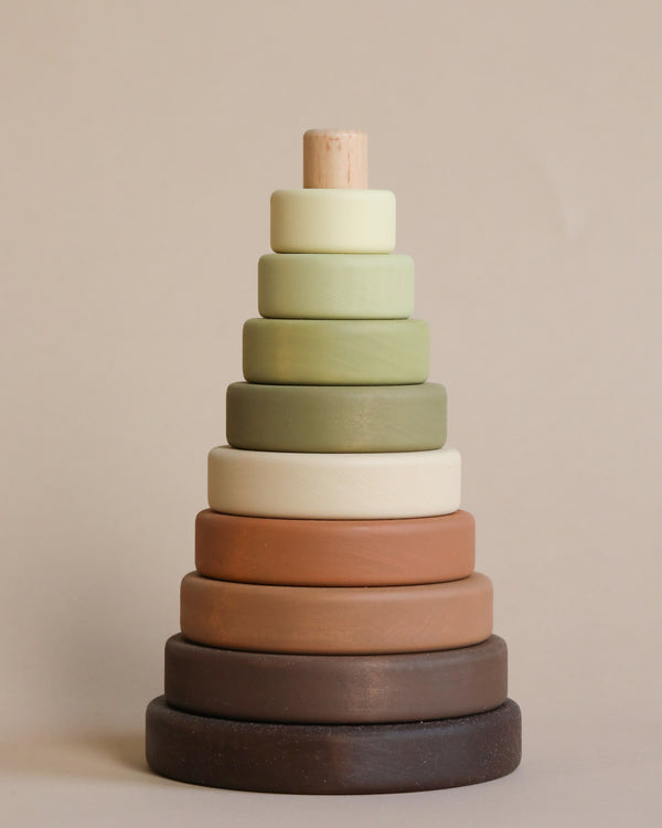 A stack of seven round, Olive wooden rings painted with non-toxic paint, in a gradient of colors ranging from dark brown at the base to light cream at the top, resembling a toddler's stacking toy.
