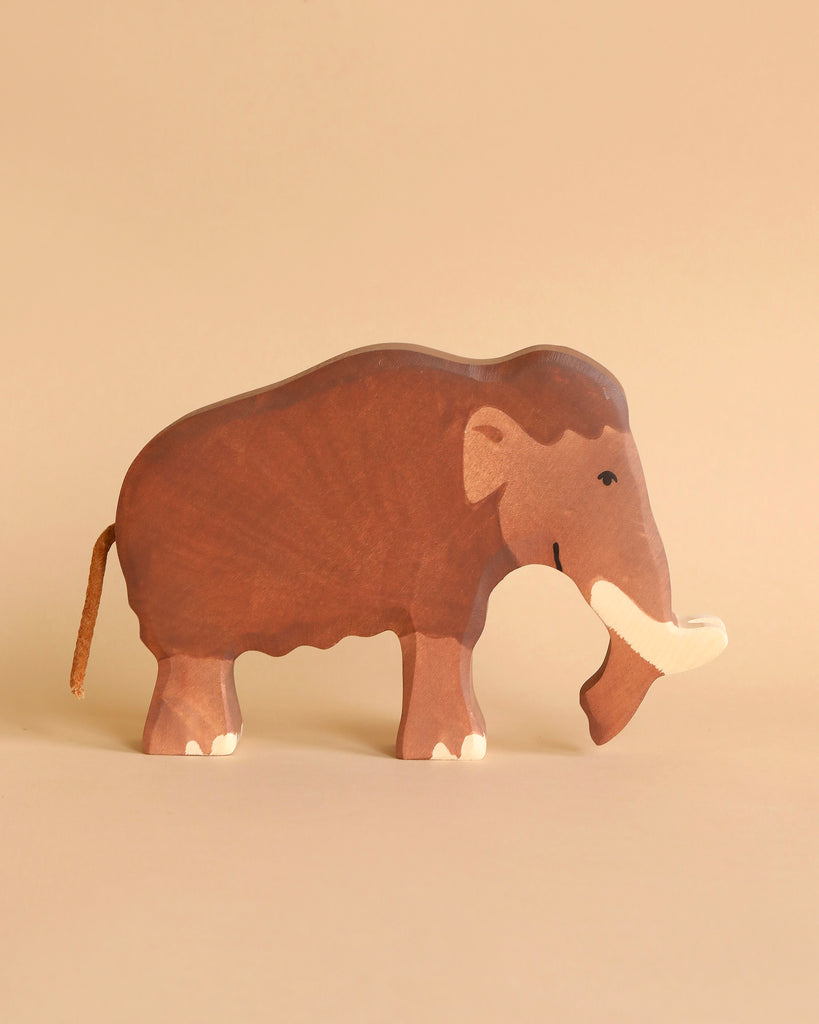 A handcrafted wooden toy Holztiger Mammoth with a dark brown body, tusks, and detailing that represent its features. Made in Europe as part of the HOLZTIGER figures collection, the Holztiger Mammoth stands against a matching beige background.