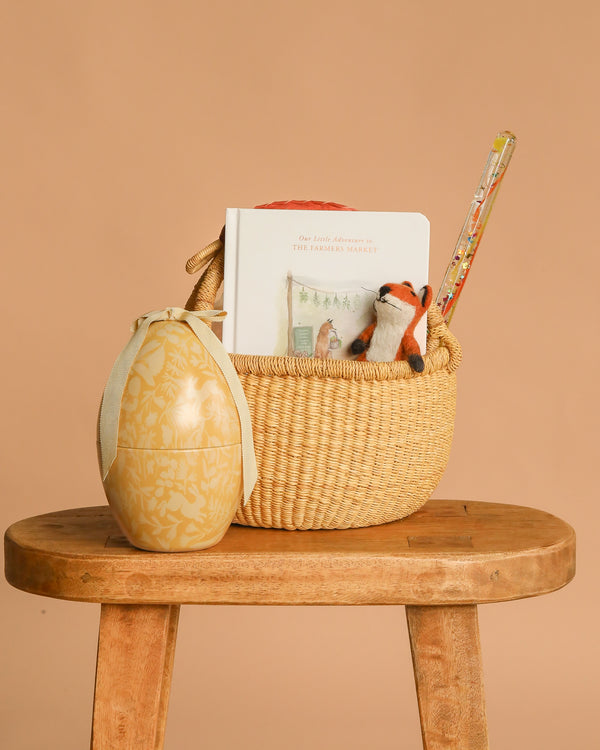 The Easter Basket Gift Set, featuring a Farmers Market board book, plush fox toy, and pencil in a basket on a wooden stool beside a Maileg Easter Egg, is displayed against a beige background.
