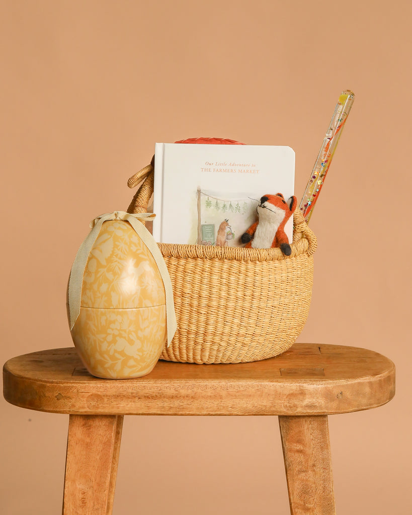 The Easter Basket Gift Set, featuring a Farmers Market board book, plush fox toy, and pencil in a basket on a wooden stool beside a Maileg Easter Egg, is displayed against a beige background.