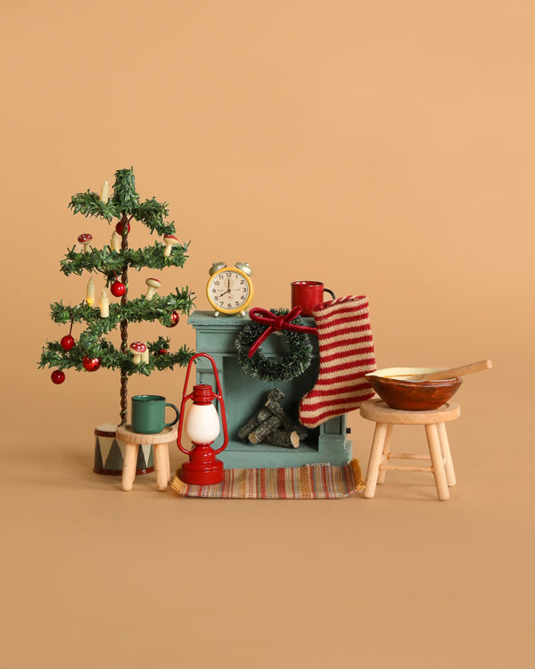 The Maileg Cozy Christmas Living Room Set features a mouse-sized Christmas tree, striped stocking, tiny fireplace, cups, lamp, clock, stools, and bowl—all arranged on a beige background for a festive miniature holiday scene.