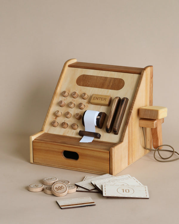 Wooden Cash Register: A sustainably sourced Wooden Cash Register with round buttons, a receipt roll, and play money, including coins and bills, set against a plain beige background.