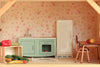 The Maileg Fully Furnished Farmhouse Set + Surprise Gift is a miniature vintage-style kitchen, perfect for a Maileg Farmhouse Dollhouse. It features a green sink, white fridge, small table with chairs, assorted toy vegetables on a wooden shelf, and charming dollhouse accessories—all set against floral wallpaper.