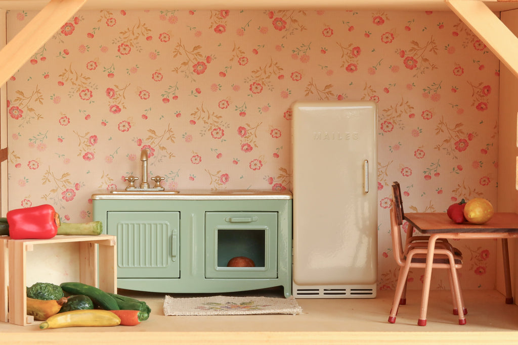 The Maileg Fully Furnished Farmhouse Set + Surprise Gift is a miniature vintage-style kitchen, perfect for a Maileg Farmhouse Dollhouse. It features a green sink, white fridge, small table with chairs, assorted toy vegetables on a wooden shelf, and charming dollhouse accessories—all set against floral wallpaper.