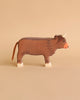 A cow from the Holztiger Handmade Wooden Farm Animals Set, crafted from maple and beech wood, features a brown body, white hooves and horns, and a brown tail. It stands on a beige surface against a beige background.
