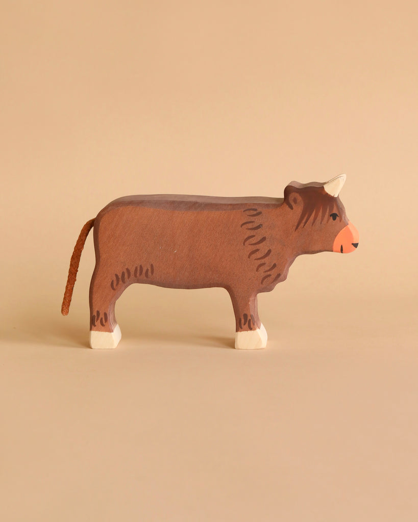 A cow from the Holztiger Handmade Wooden Farm Animals Set, crafted from maple and beech wood, features a brown body, white hooves and horns, and a brown tail. It stands on a beige surface against a beige background.