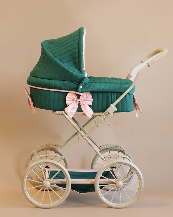 Introducing the Doll Pram - Bowie Smoke Pine: This pram showcases a charming vintage design made from recycled materials. It features a quilted green cover adorned with pink bows, complemented by a sturdy white frame, large wheels, and a beige handle. All of this is set against a soft beige background, effortlessly combining classic elegance with eco-conscious appeal.