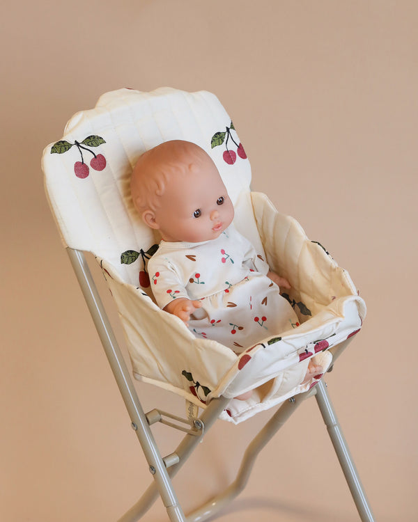 A Konges Sloejd High Chair in Cherry Glitter fabric, featuring a doll dressed in a matching cherry-patterned outfit, is set against a beige background. This charming setup, enhanced with delightful doll accessories, adds an inviting touch to any playroom.