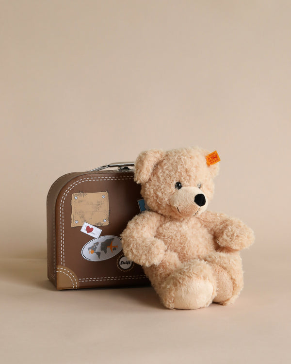 A Steiff, Fynn Teddy Bear in Suitcase with a Button in Ear sits next to a small suitcase decorated with travel stickers, against a plain beige background.