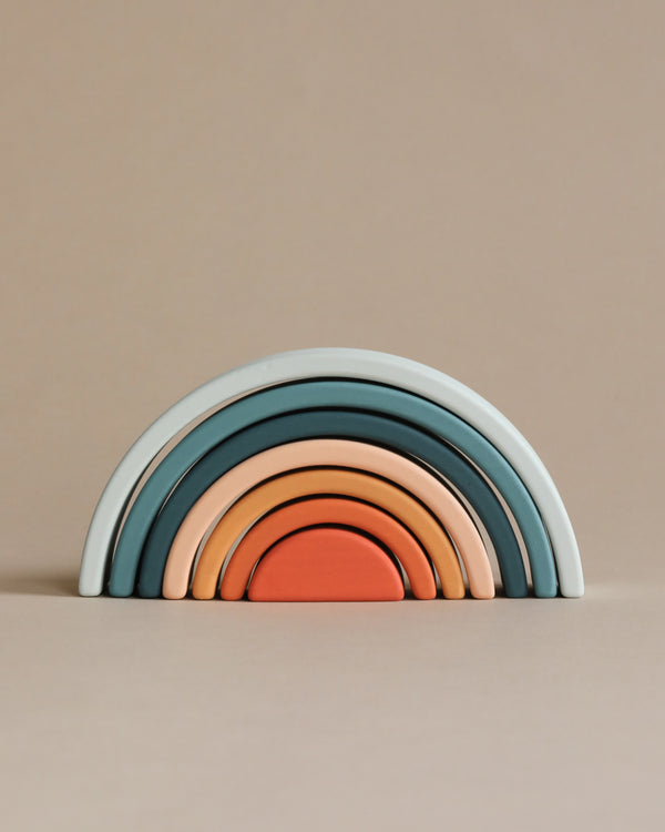 A stack of colorful, semi-circular wooden arches arranged in order of size to form a rainbow shape, designed for open-ended play and set against a neutral beige background. This handmade Mini Rainbow Stacker - Lagoon is sure to provide hours of fun!